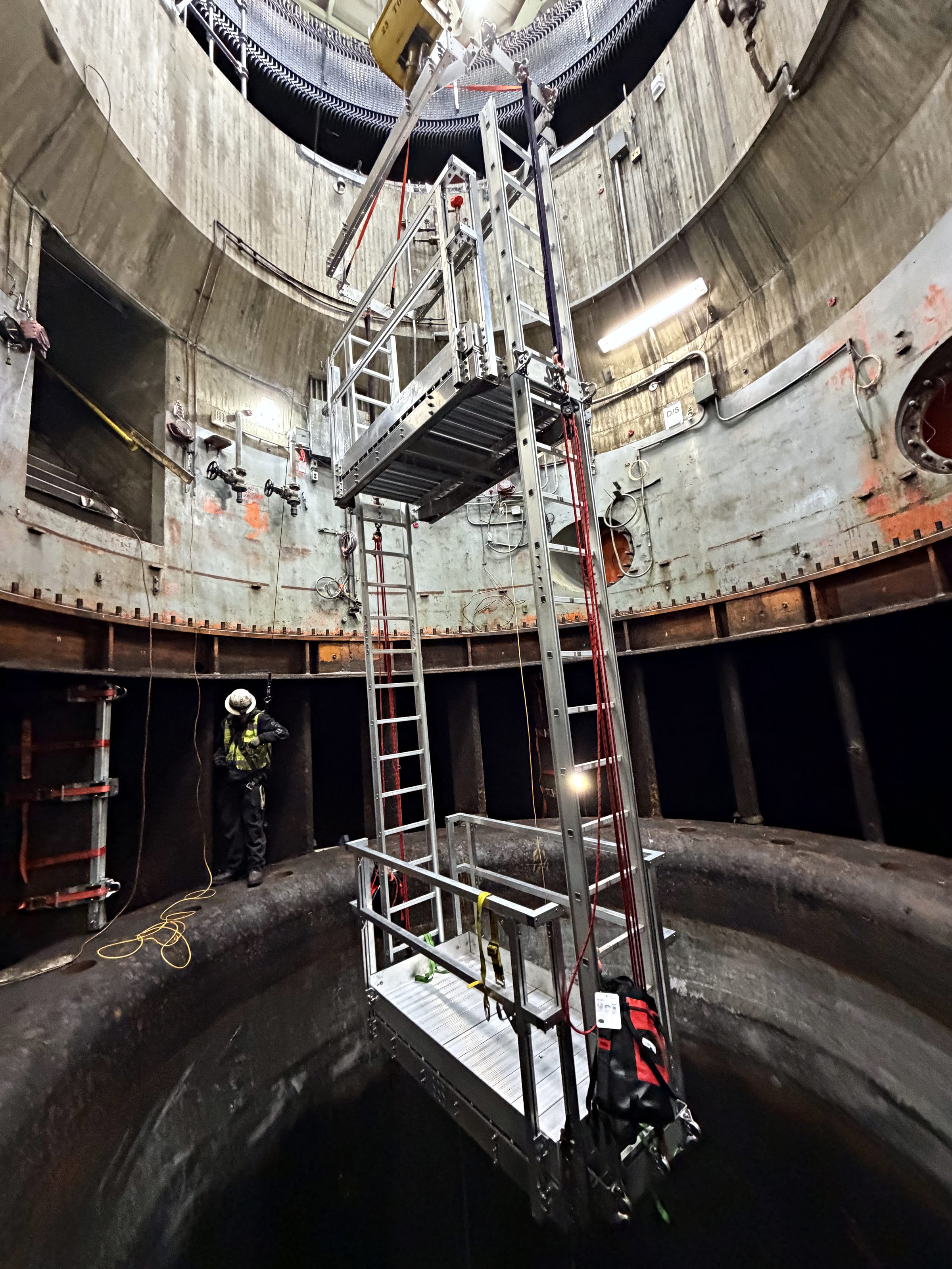 Aluminum platform and ladder assembly suspended from a crane inside a large steel-lined shaft, with a worker standing below.