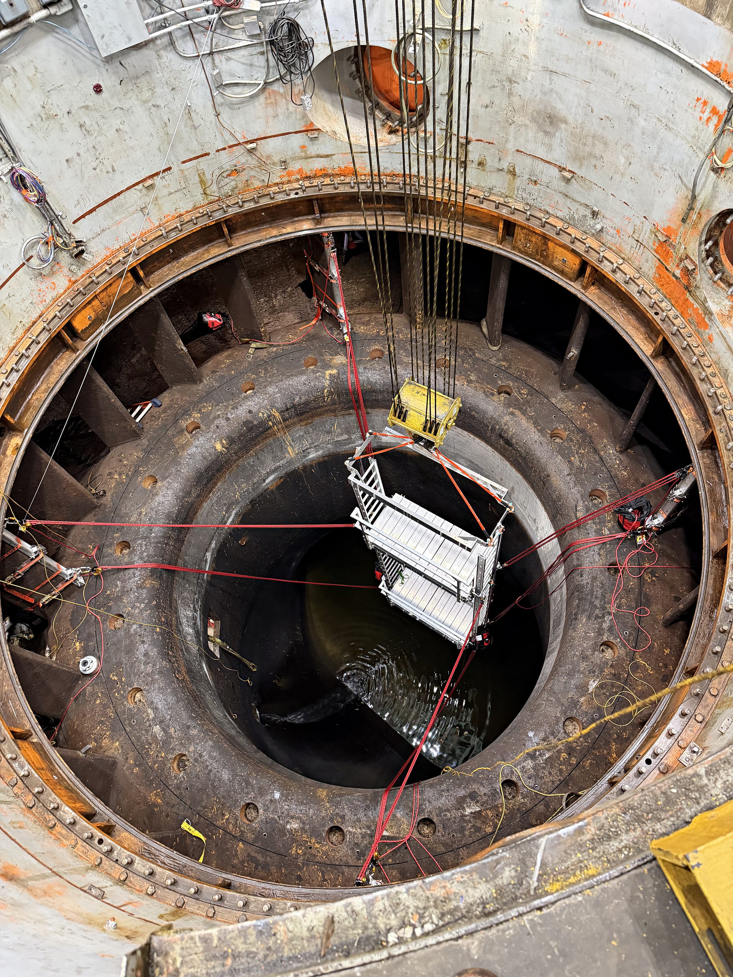 Looking straight down a steel-lined shaft at an aluminum platform and ladder assembly suspended from a crane.